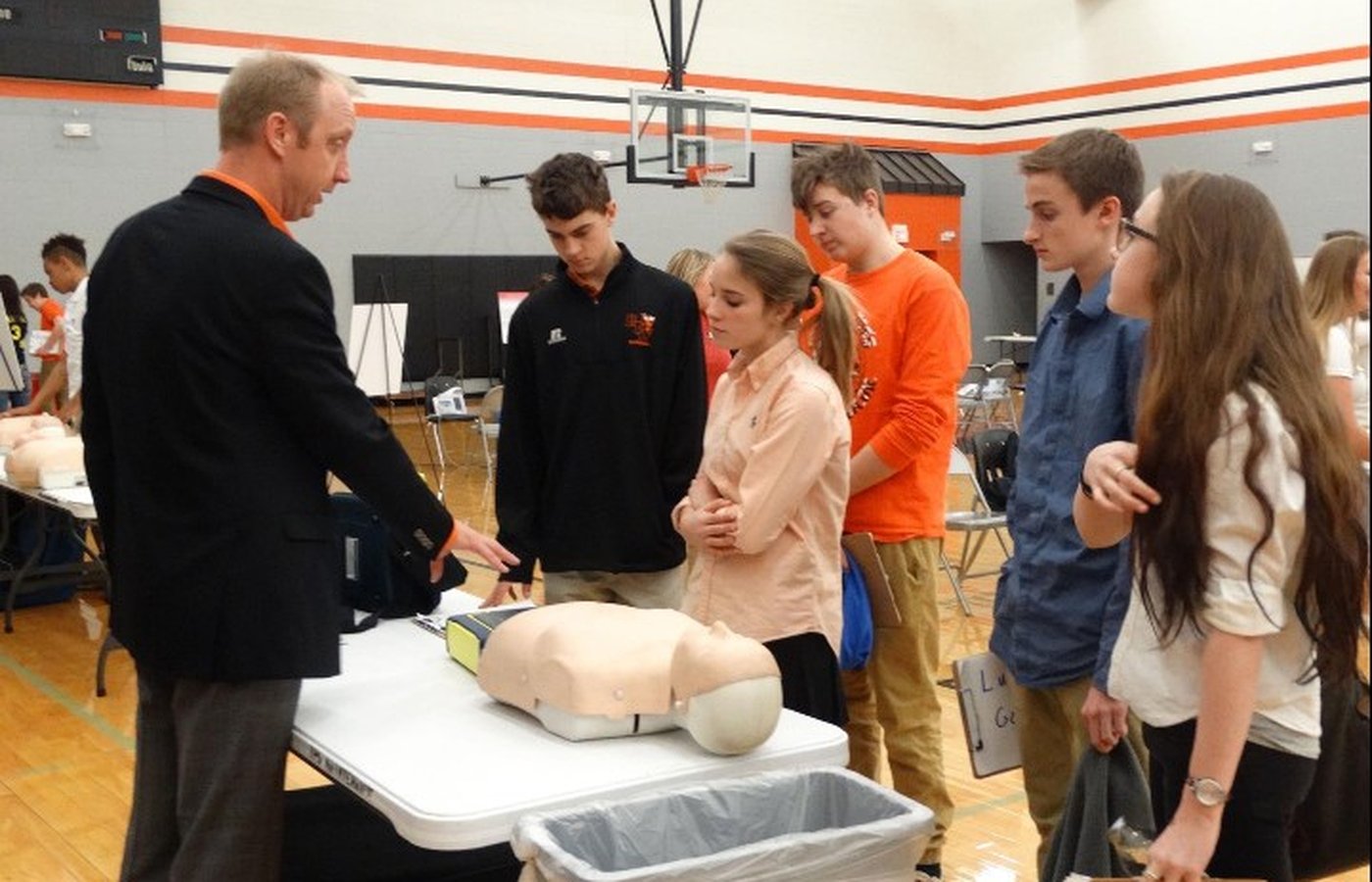 A foundation volunteer in a dark blazer demonstrates CPR technique using a manikin and an automated external defibrillator (AED) to a group of high school students gathered around a training table in a gymnasium