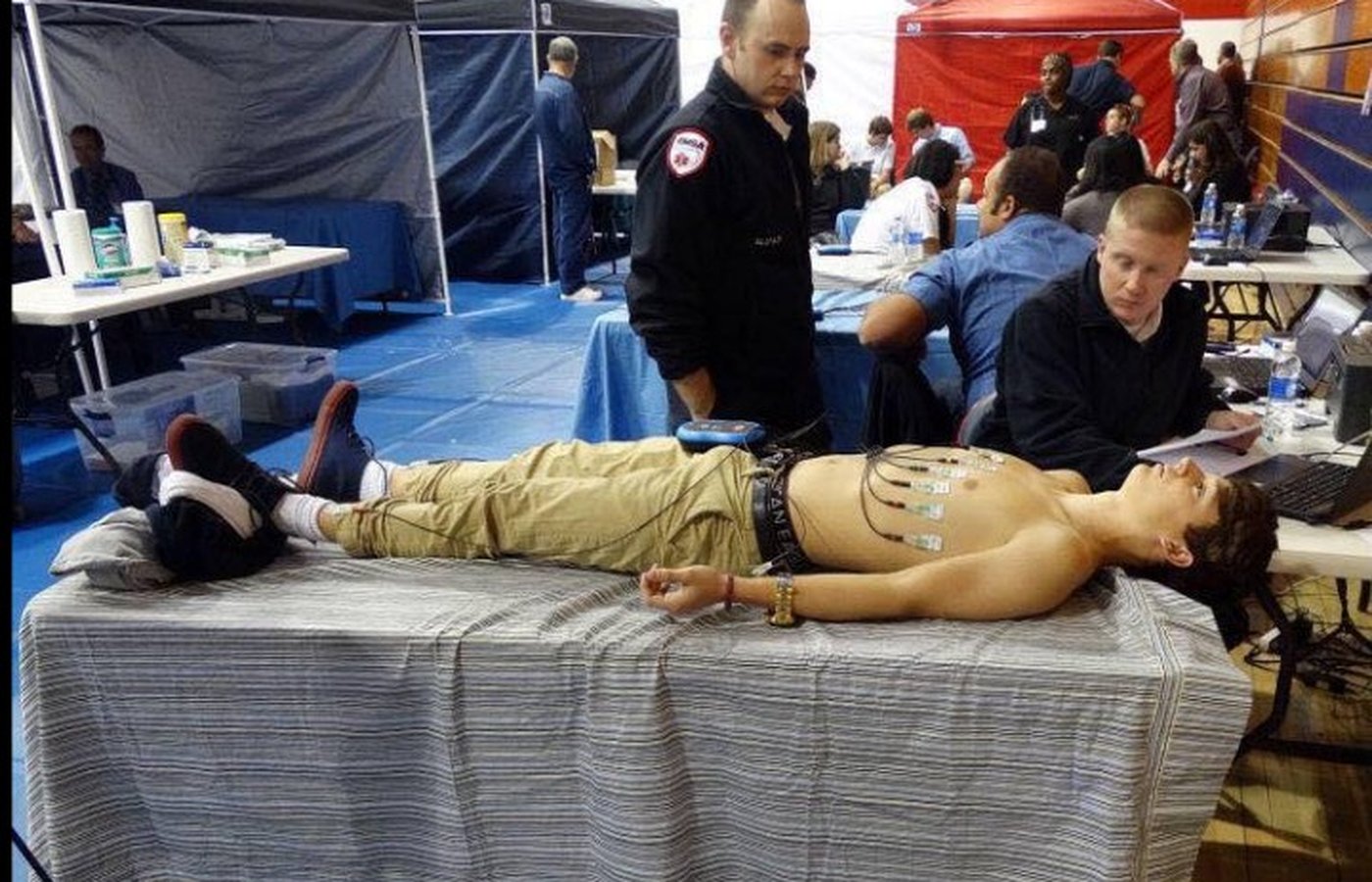 A student receives a 12-lead electrocardiogram at a foundation screening event. An emergency medical technician stands beside the table, and a technician at right reviews the tracing on a laptop.