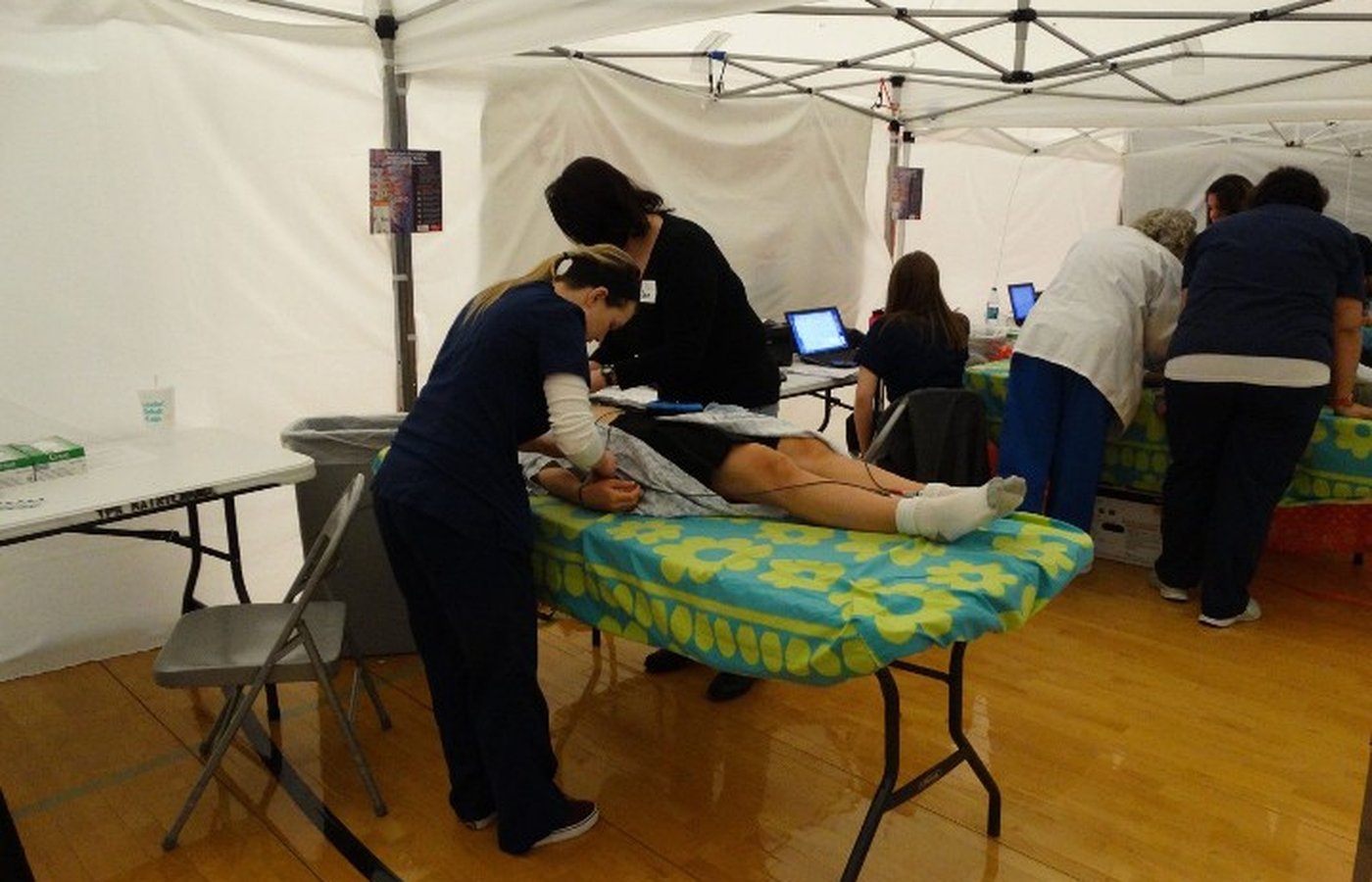 A sonographer in scrubs performs an echocardiogram on a student at a foundation screening event under a privacy tent, with additional examination stations visible in the background