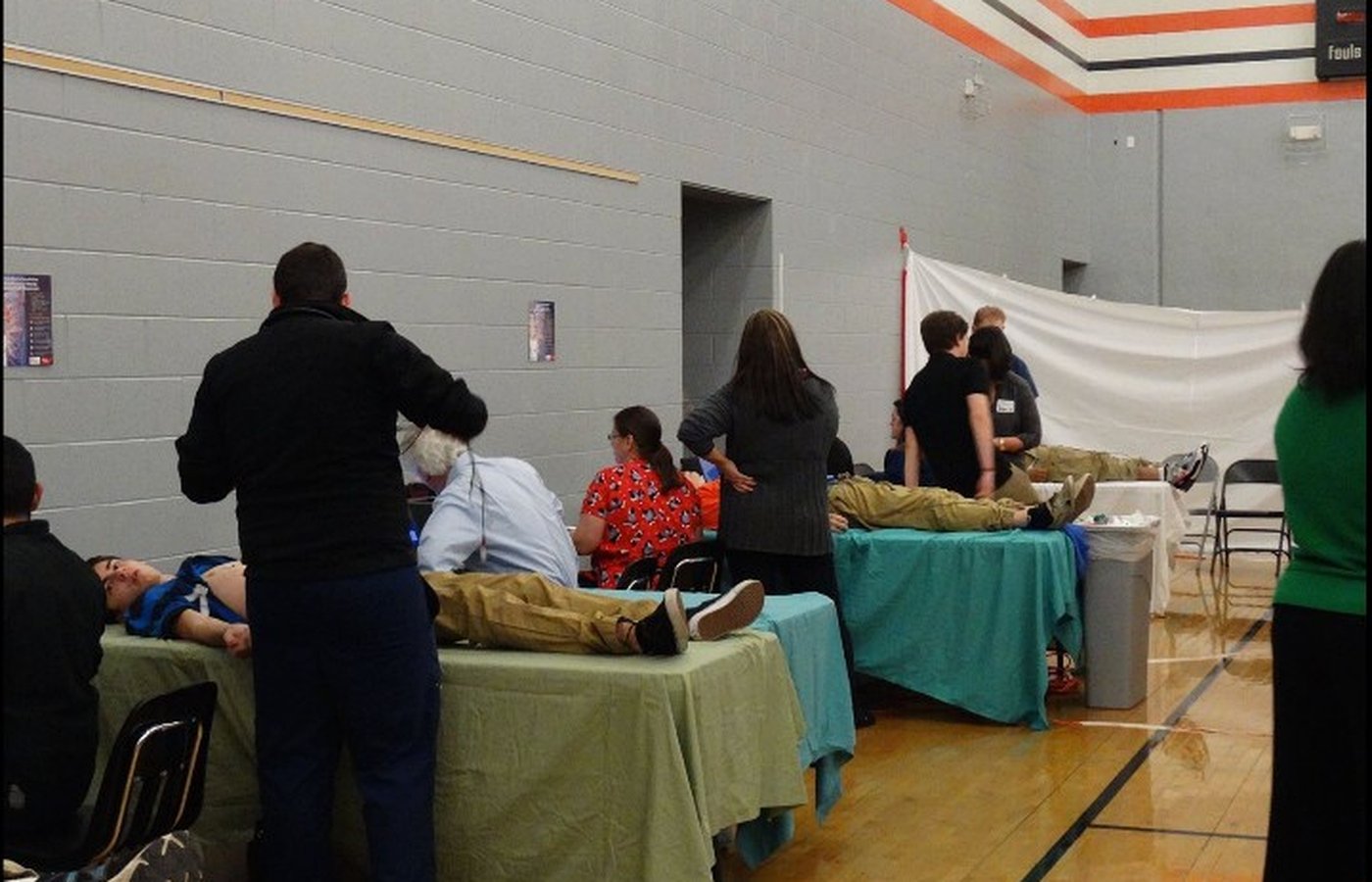 A wide view of a foundation screening event in a school gymnasium, showing multiple ECG stations running in parallel with students on examination tables and medical volunteers in scrubs